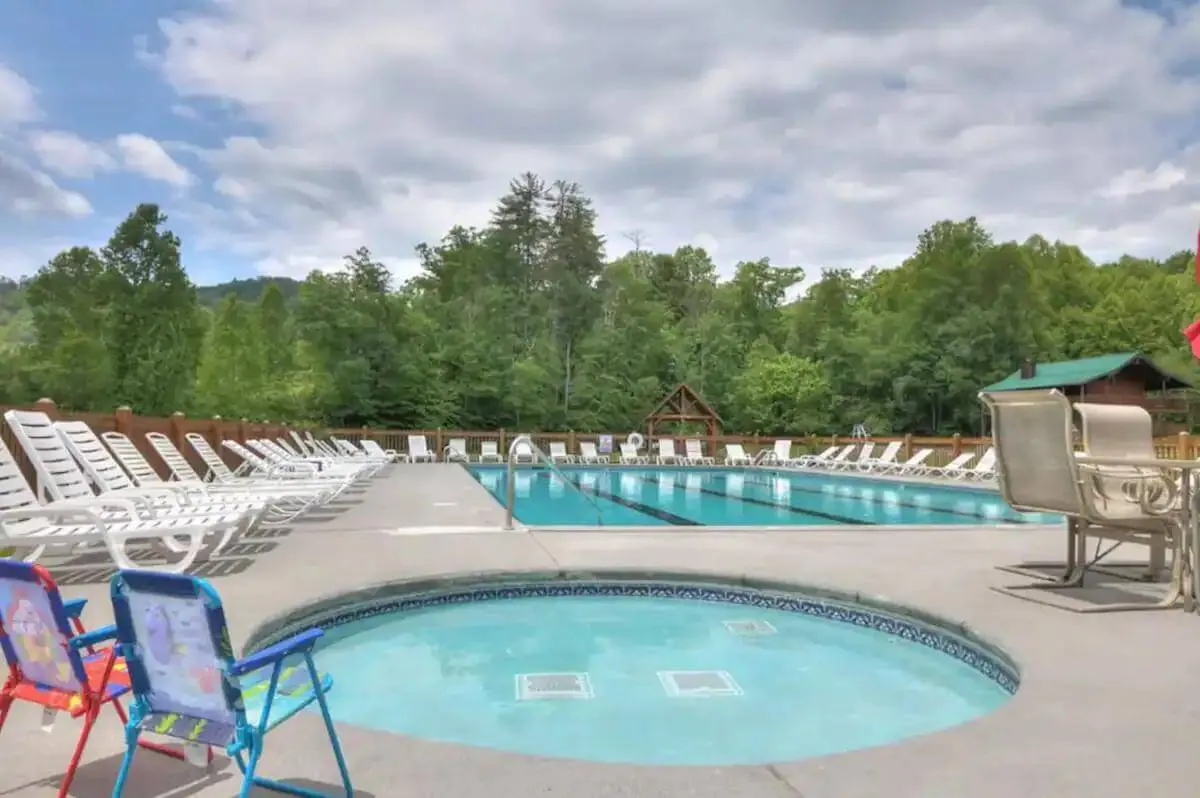 A sparkling swimming pool and jacuzzi are surrounded by comfortable lounge chairs and set against a backdrop of lush trees and distant mountain views. The scene is perfect for family gatherings, relaxation, and enjoying sunset views over the mountains.