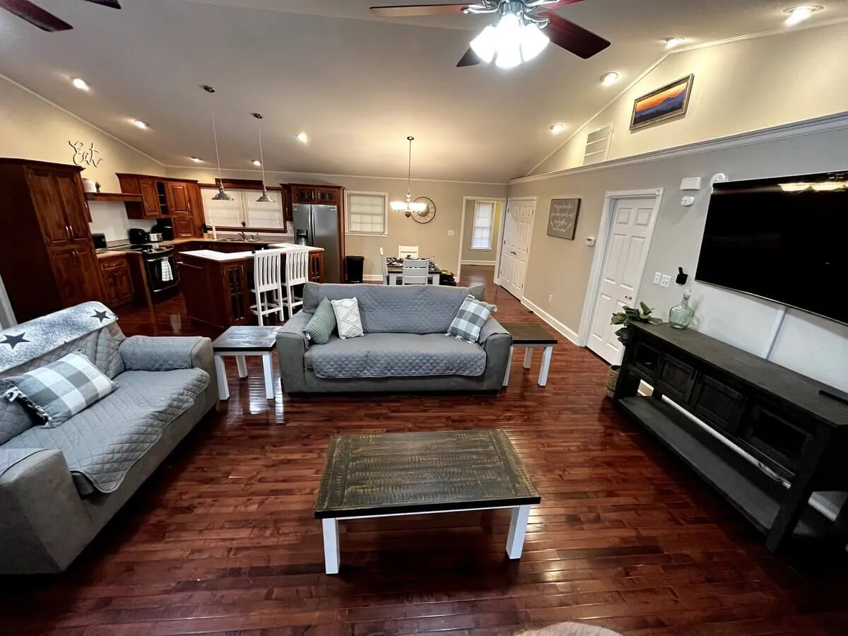 An open-concept living area with comfortable sofas, a central coffee table, and a kitchen space in the background. Large windows allow natural light to stream in, creating a bright and airy atmosphere ideal for group gatherings.
