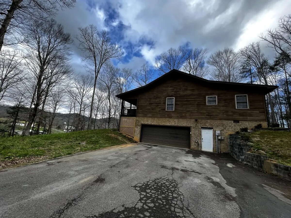 This image features the exterior of a wooden house set among tall trees, with a garage door visible and the ambiance of a peaceful retreat. The setting highlights the connection to nature with outdoor spaces ideal for exploration or relaxing after a trail walk.