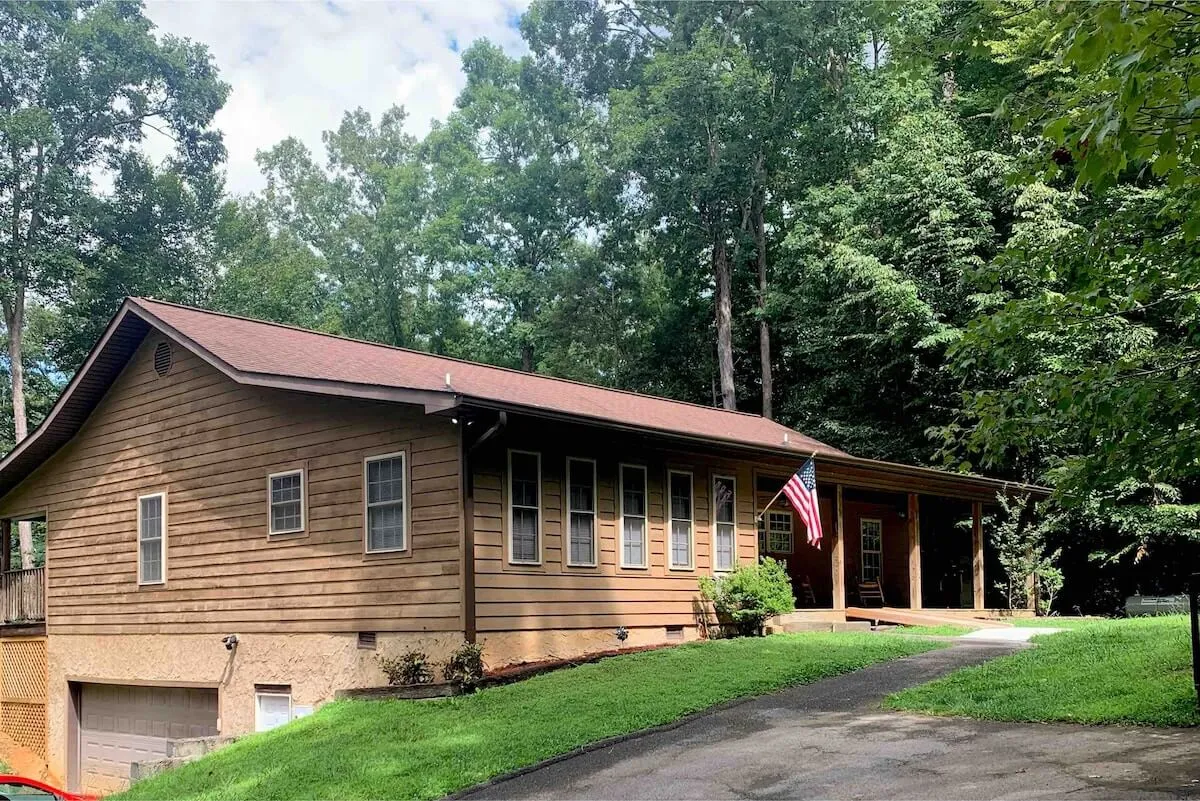 The image showcases a charming wooden cabin nestled among tall trees, with a welcoming front porch adorned with an American flag. The porch invites guests to relax and take in the peaceful woodland surroundings, setting a tranquil, inviting mood.
