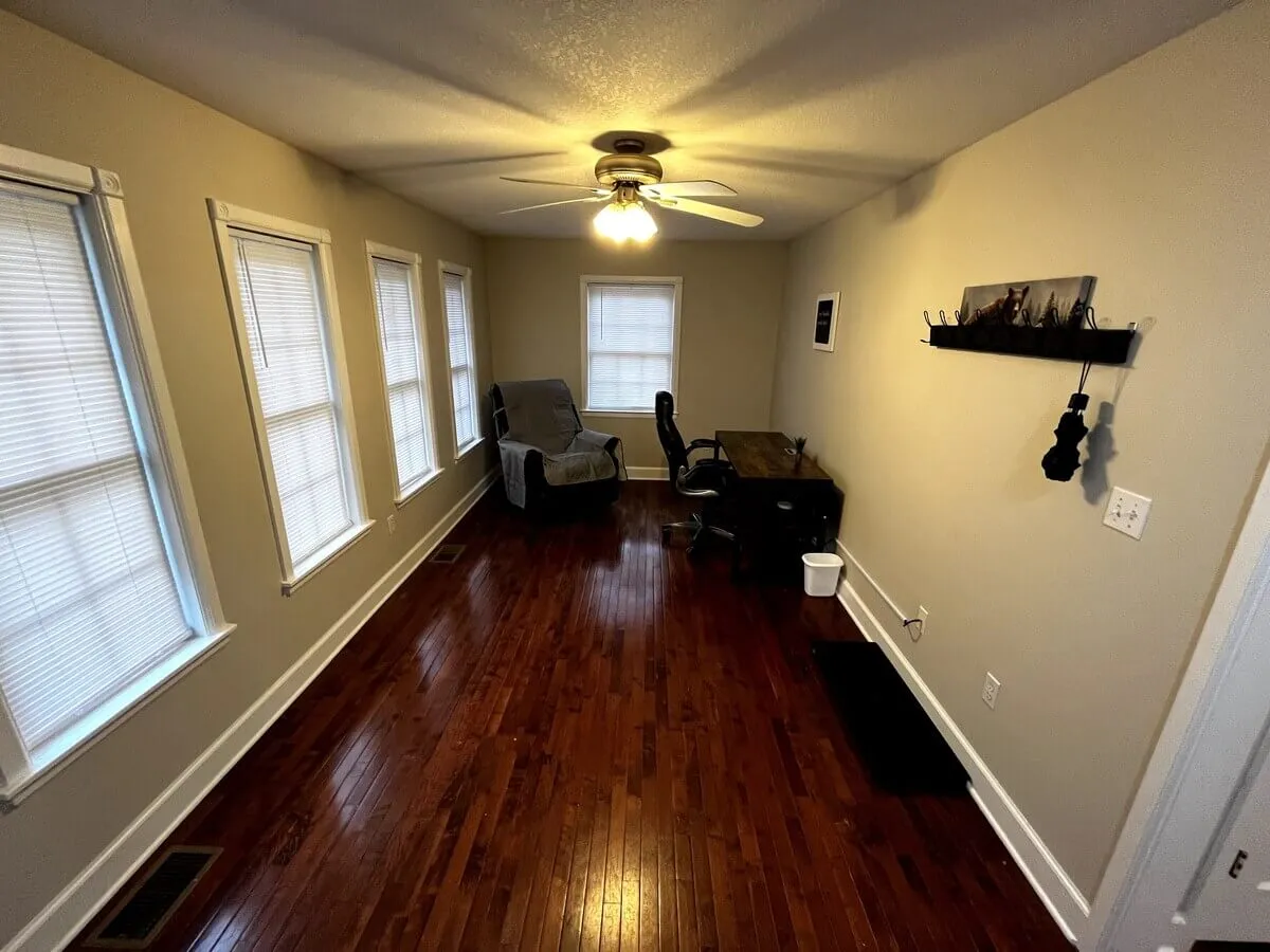 A peaceful nook featuring a desk with a chair, a recliner, multiple windows letting in natural light, and a wooden floor. The space is styled for reading, relaxing, or quiet conversation, with cozy and functional touches.