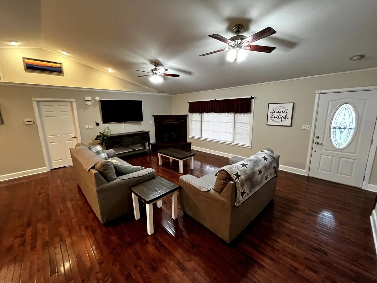 A spacious and inviting living room featuring a plush sofa set arranged around a coffee table and a cozy fireplace, with a television and the front door visible. The space is warmly lit, showcasing ample room for family gatherings and socializing.