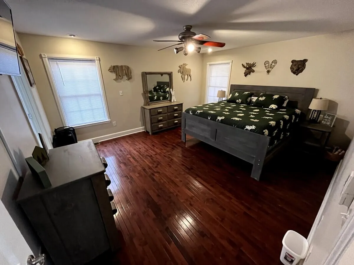 A cozy bedroom featuring a bed with patterned bedding, a wooden dresser, a mirror, and decorative animal heads on the wall. Sunlight streams through windows, creating a warm, rustic atmosphere ideal for sleeping, reading, or unwinding.