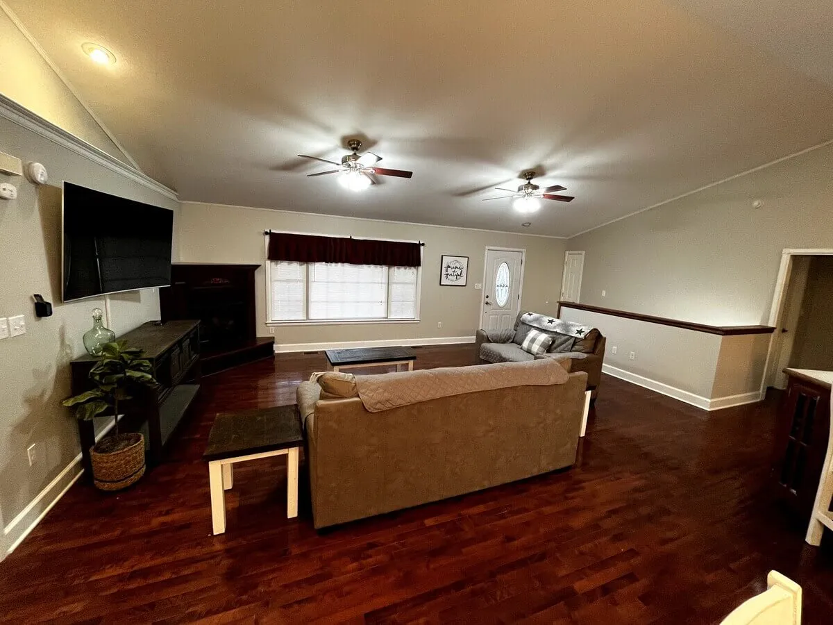 A cozy corner of the living room with a soft sofa, coffee table, large window, and television. The natural light pouring in from the window adds to the welcoming, relaxed mood of the space.