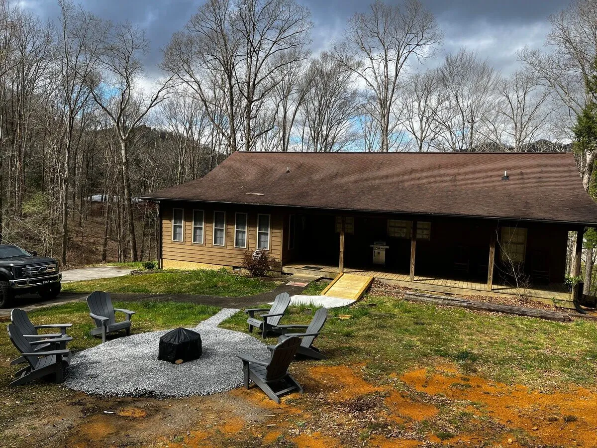 This image showcases the wooden exterior of the home nestled among mature trees, with a welcoming fire pit surrounded by chairs, set against the backdrop of the Smoky Mountains at sunset. The setup is ideal for campfire gatherings and outdoor relaxation, offering a cozy, communal ambiance for groups to socialize under the open sky.
